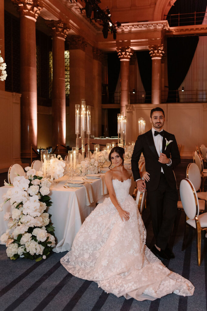 Bride and groom posing in a columned ballroom beside a long candlelit table and tall white floral arrangements.
