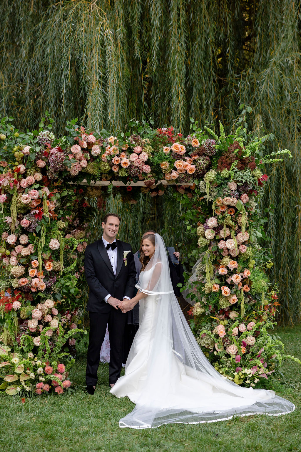 Couple holding hands at the altar under a lush floral arch of peach and pink roses with trailing greenery, on a garden lawn.