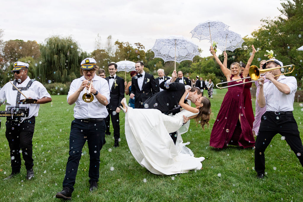 Groom dipping the bride on a lawn while a brass band in white uniforms plays and guests hold white parasols.